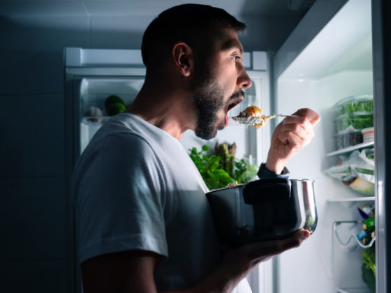Ein Mann steht im Dunklen vor dem geöffneten Kühlschrank und isst etwas.